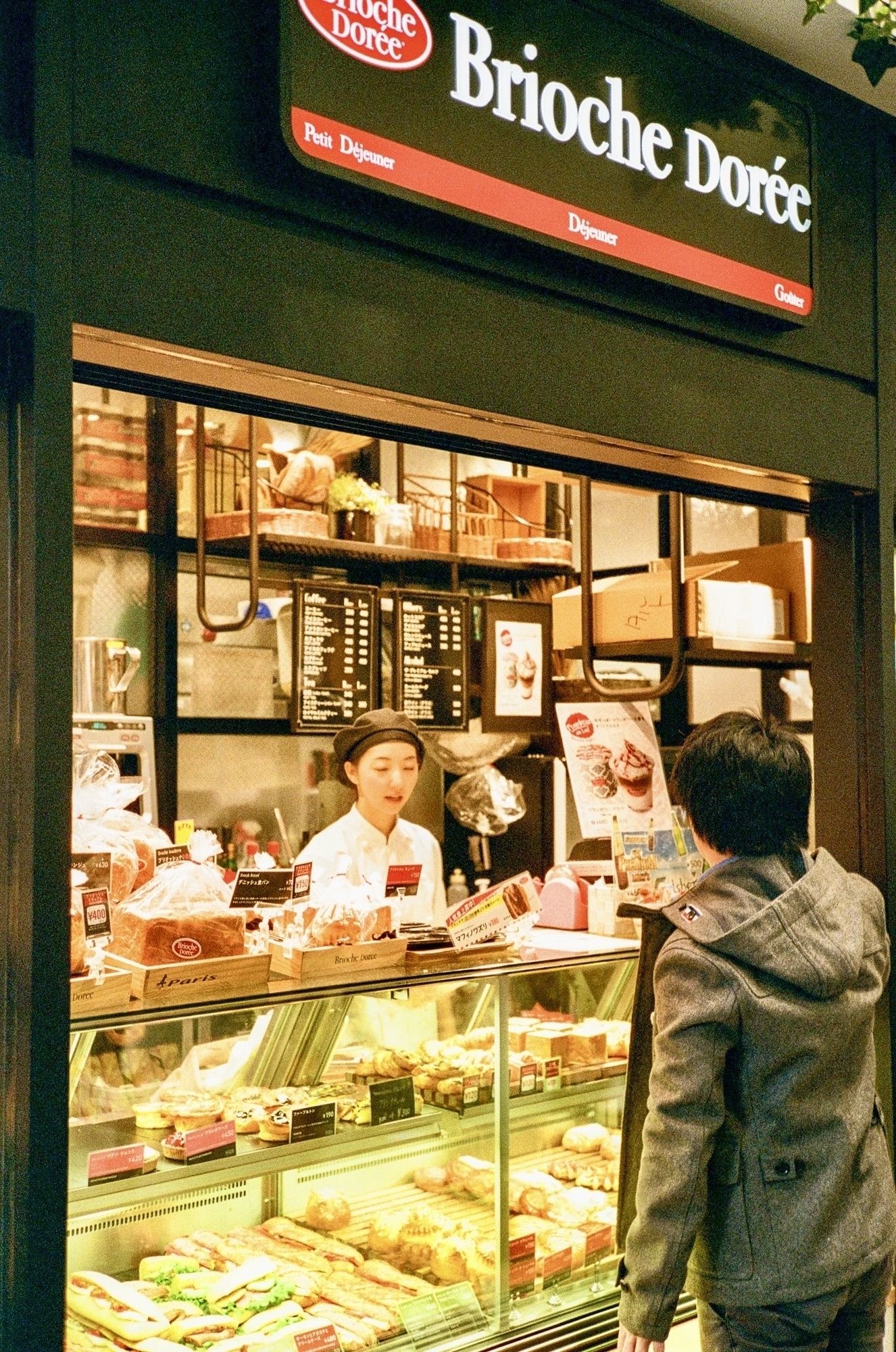 Young woman working a bread shop counter in the evening, with a customer in a grey-green hoodie making a purchase. The light is warm and inviting. 