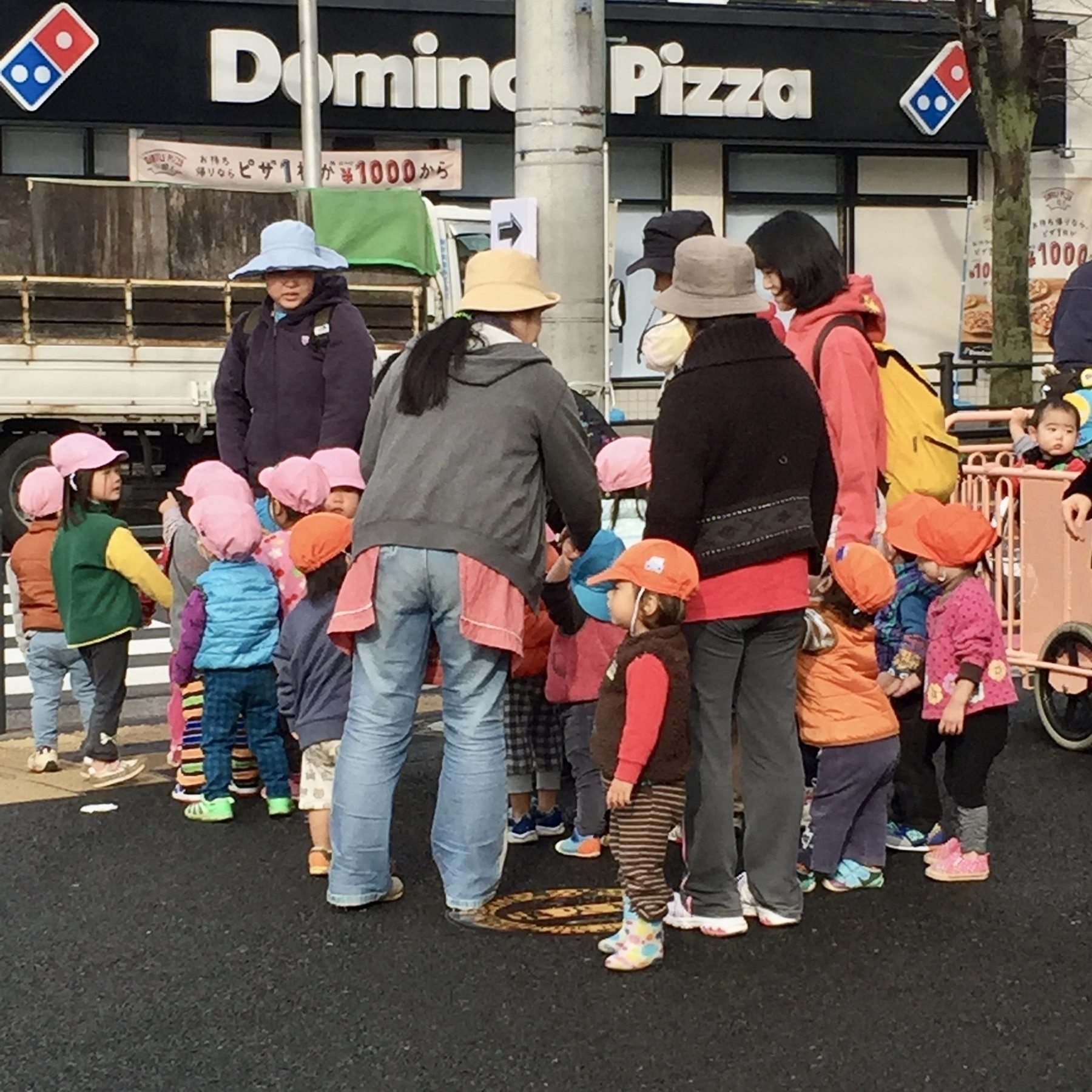 Kindergarten teachers surrounding their students at a traffic stop light in Yokohama Japan. The children are wearing various colored hats indicating their class. 