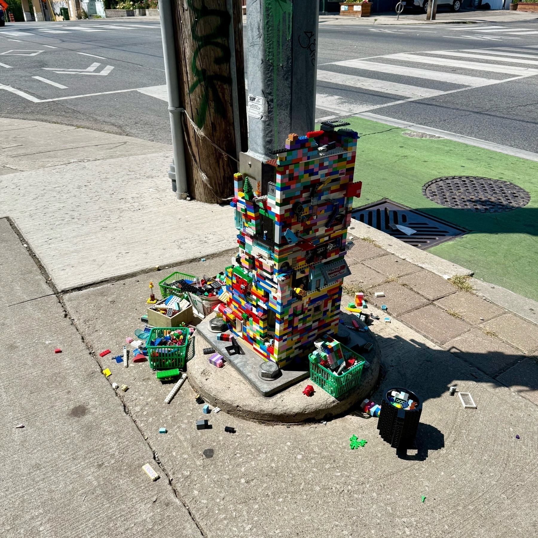 A traffic light pole base on a city sidewalk is covered in colorful LEGO bricks, forming a tall, blocky structure. Small baskets filled with loose LEGO pieces surround the base, and scattered bricks are spread across the pavement.