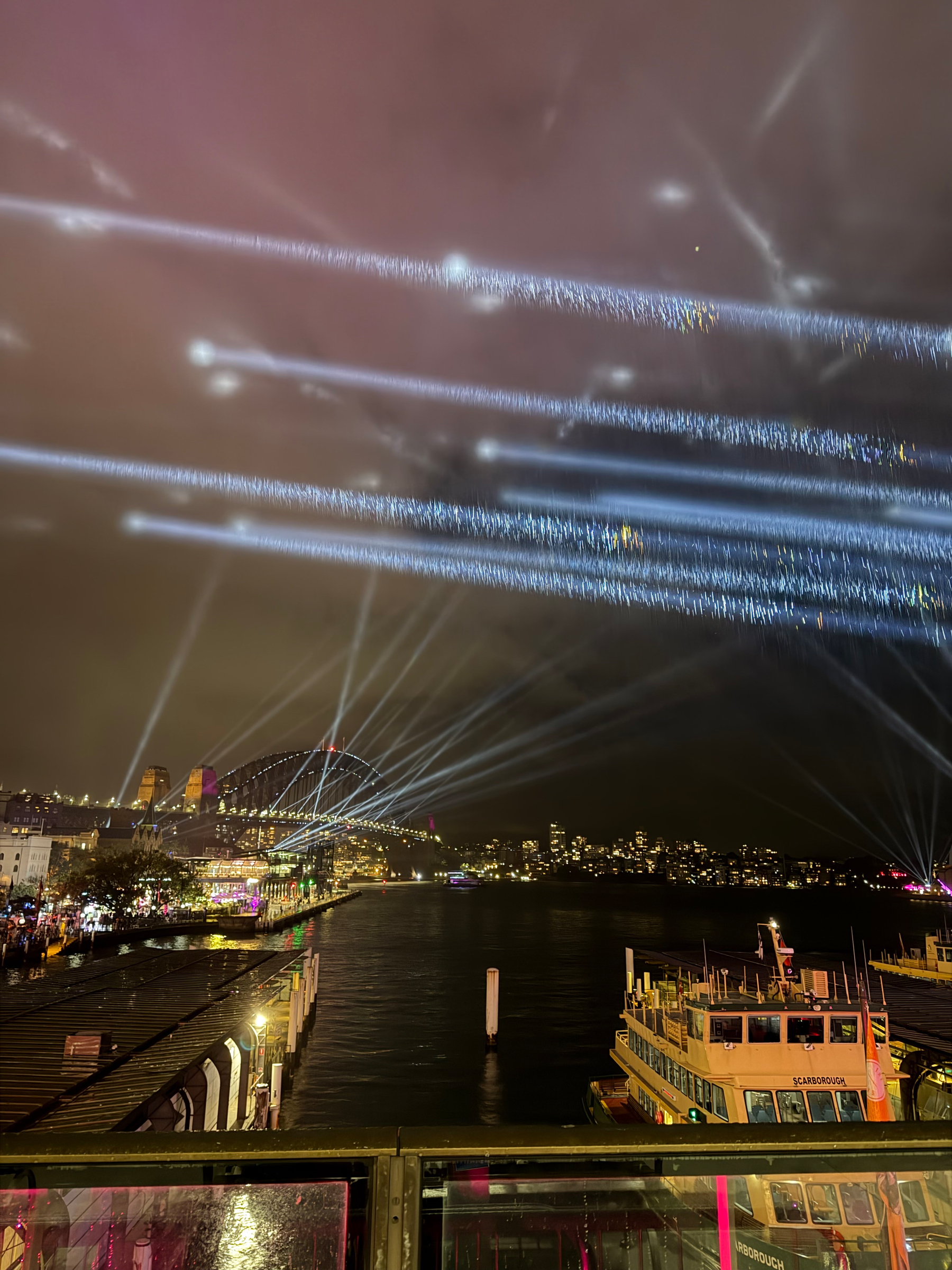 Spotlights illuminate the night sky over Sydney harbor with boats and a lit-up bridge in the distance.