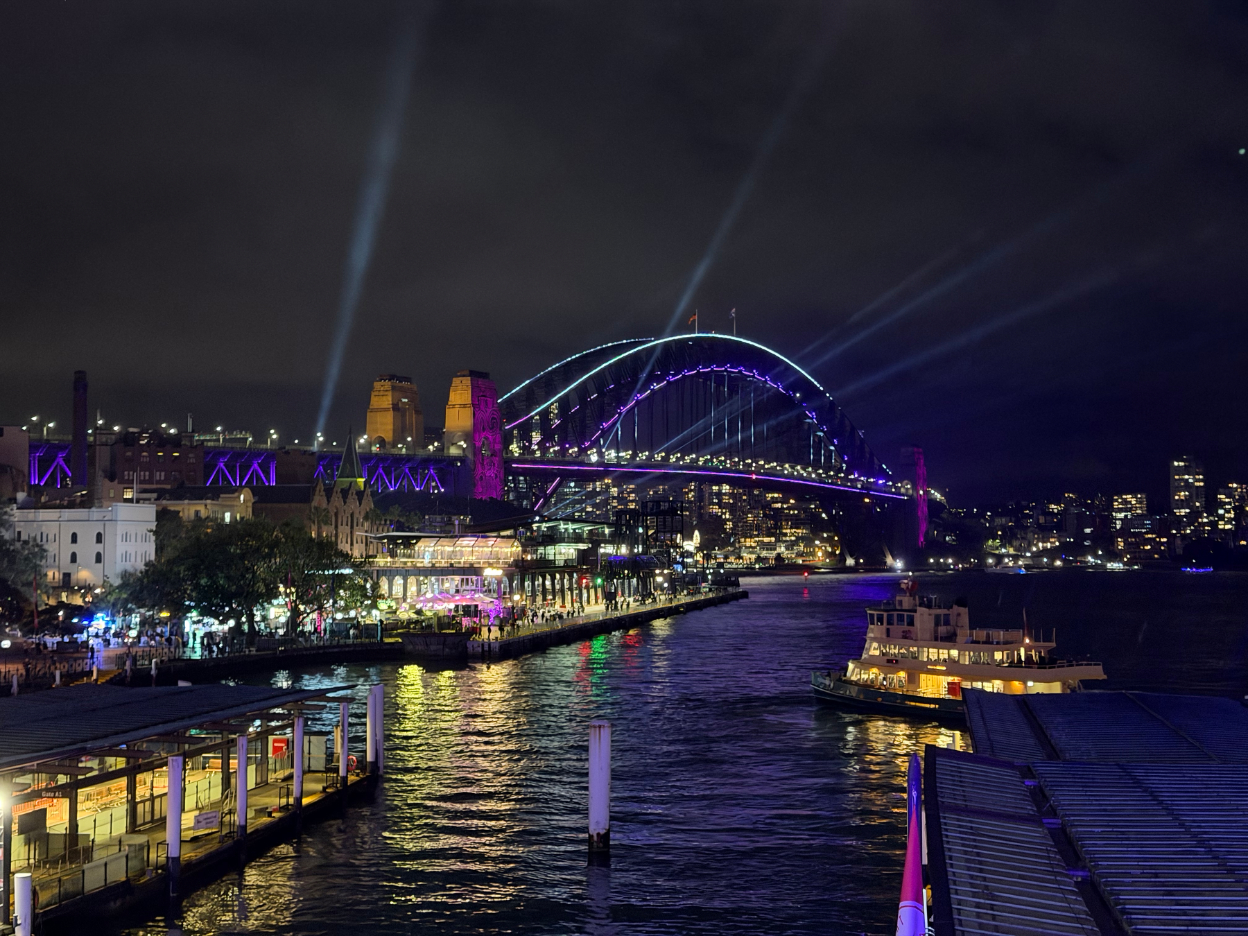 A brightly illuminated Sydney Harbour Bridge is set against a night sky, with vibrant lights and a lit-up ferry on the water below.