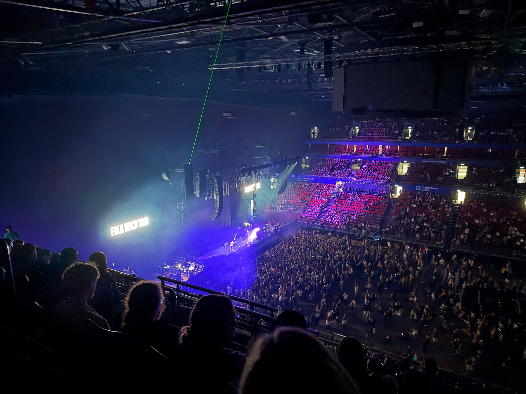 An indoor concert arena is filled with people, with a lit stage and a large screen displaying text.