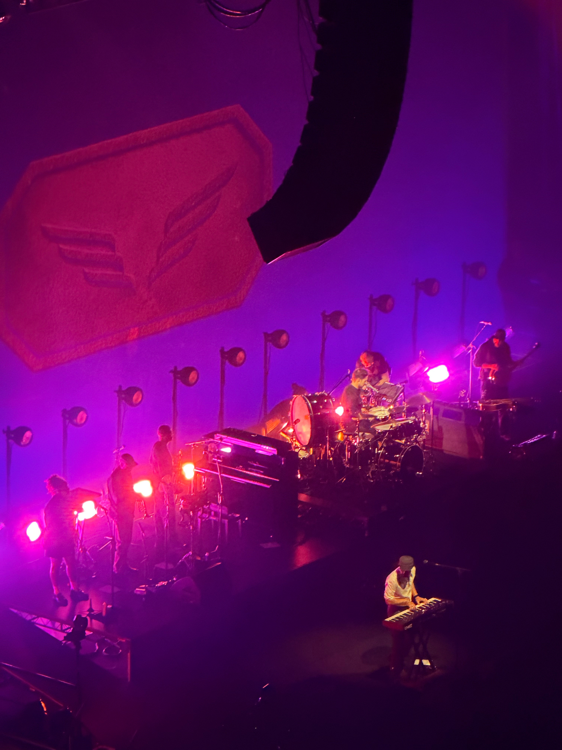 A band performs on stage under vibrant purple and pink lights with a large backdrop and multiple spotlights.