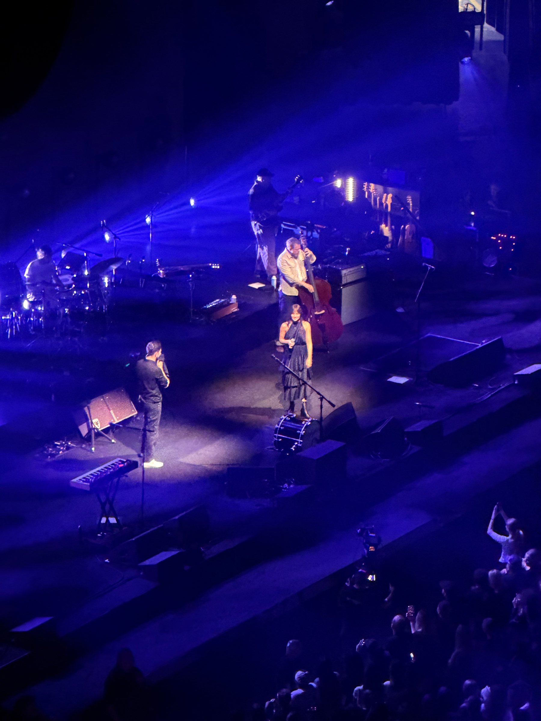 A band is performing on stage under blue and purple lights with an audience watching.