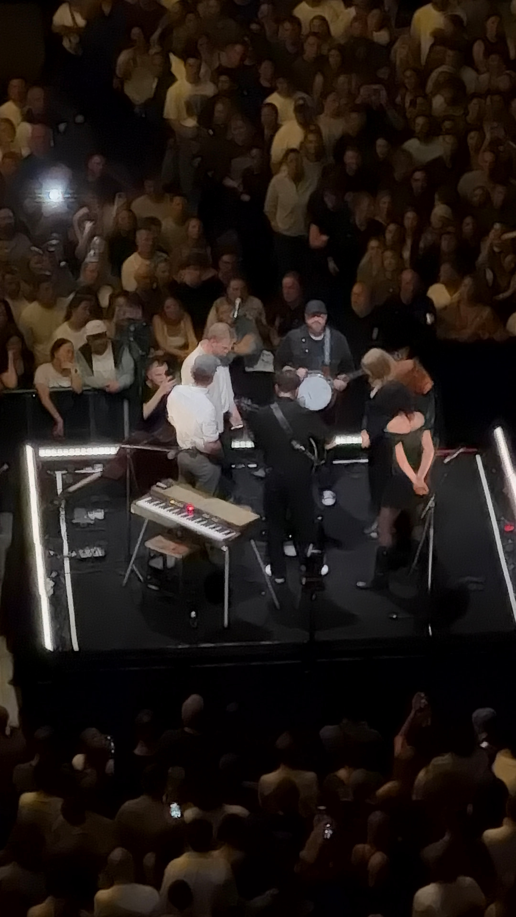 A group of musicians performs on a small stage surrounded by a large crowd in an indoor venue.