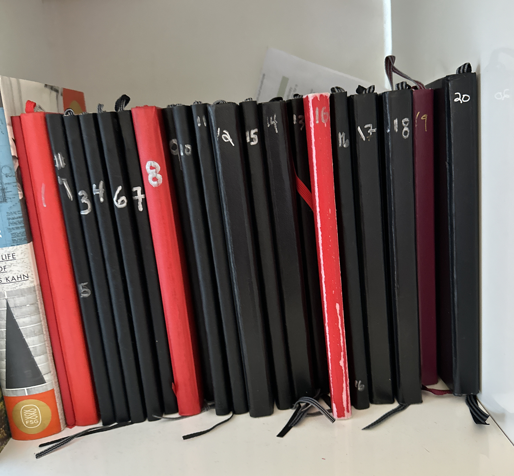 A row of mostly black and some red notebooks on a bookshelf. Each notebook spine is numbered.