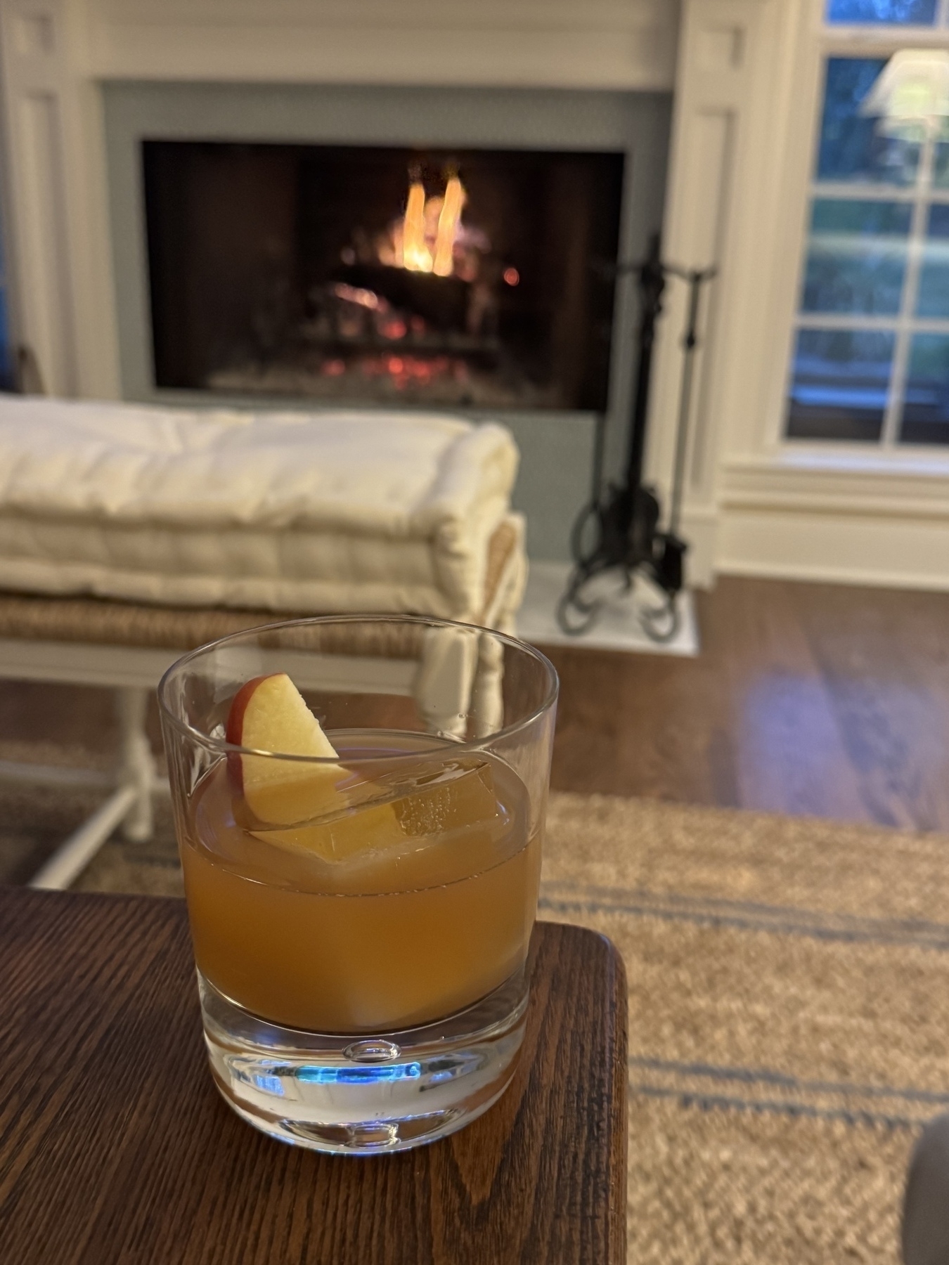 A whiskey cocktail garnished with an apple slice sits on the corner of a coffee table. A cozy fire burns in the fireplace in the background. 