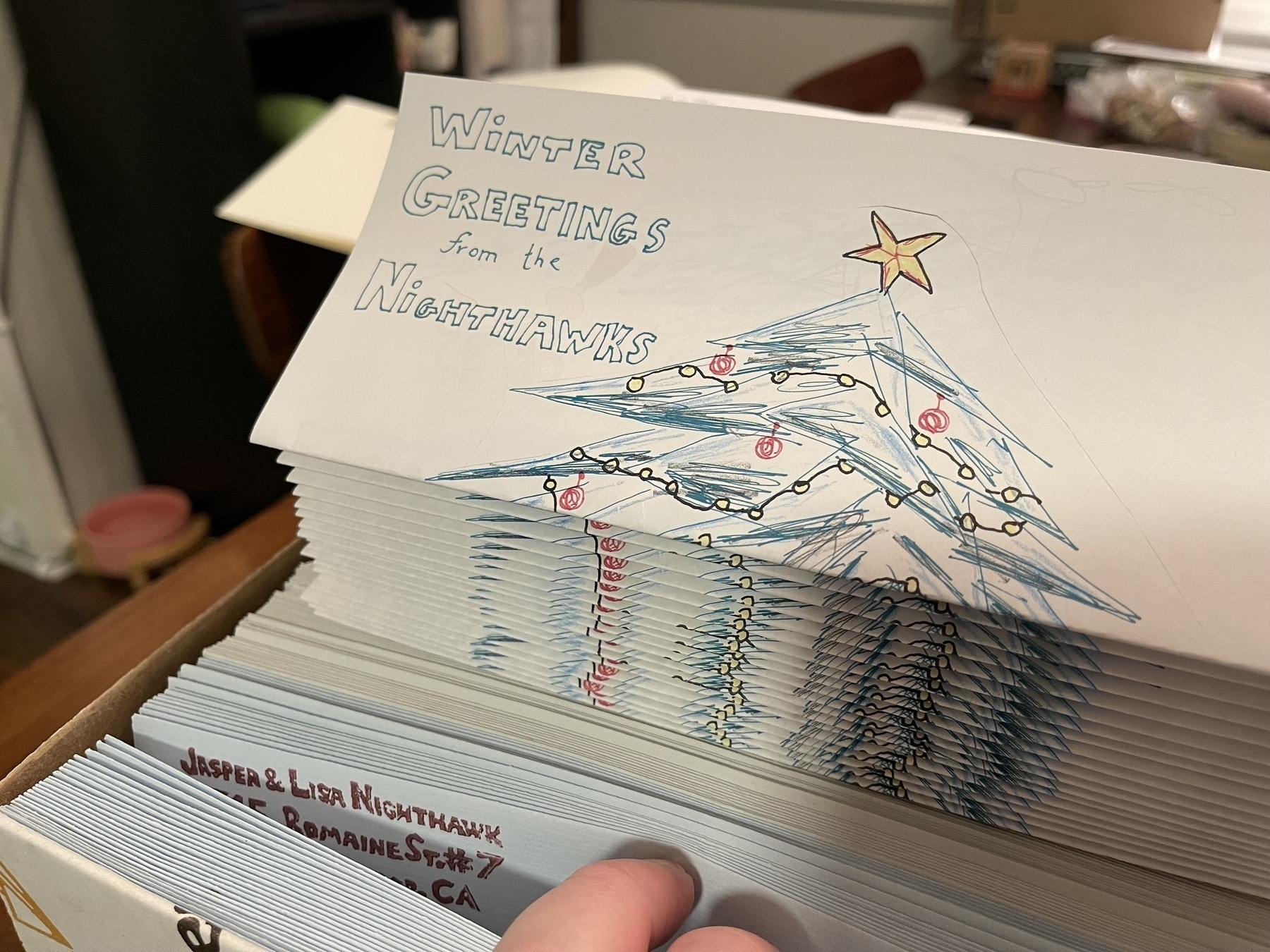 A photograph of a stack of folded letters that say “winter greetings from the nighthawks“ atop some envelopes, with a hand, barely in frame. 