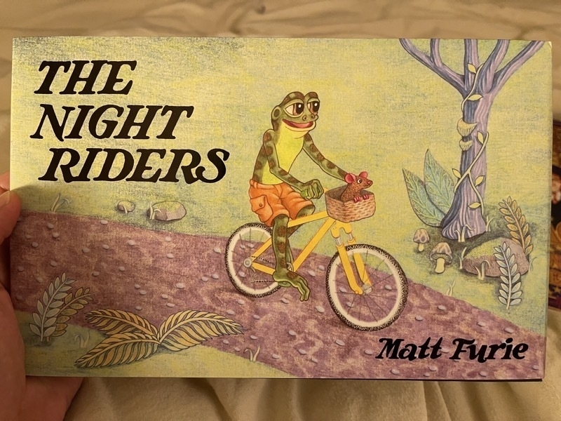 A photo of a hand holding a book. The cover shows a frog riding a bicycle and says, "The Night Riders" and "Matt Furie"