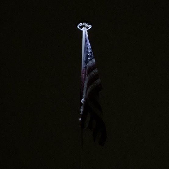 A photograph of an American flag at night. The flag is being illuminated by light camping from a narrow ring of LEDs above it.