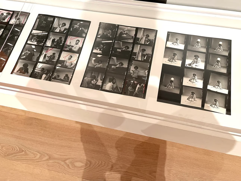 IMG_5680.jpeg.png A photo of a museum display case full of contact sheets of black-and-white photographs of Alice Coltrane with the photographer's shadow falling across the whole case