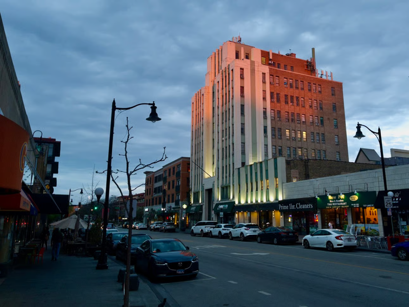 IMG_6034.jpeg.png A photo of downtown Oak Park at sunset with a tall, art deco building lit up in amber light.