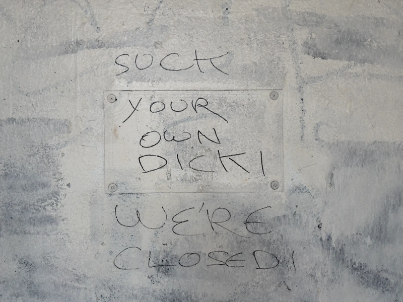 A photo of a rough scrawl of the words "SUCK YOUR OWN DICK! WE'RE CLOSED!" on uneven white paint