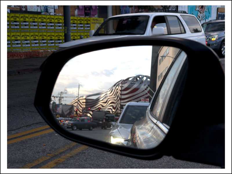 A photograph of a car's rear view mirror with a strange building in the reflections.