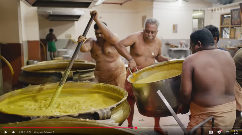 A still from a YouTube video showing two strong, shirtless men pouring a giant vat of yellow dal into a third massive vat, which is being stirred by another man.