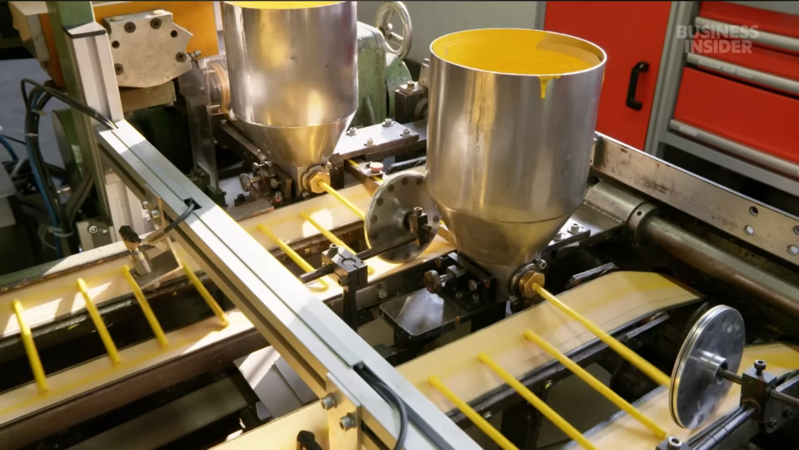 A still from a video showing yellow colored pencil cores being extruded from stainless steel vats