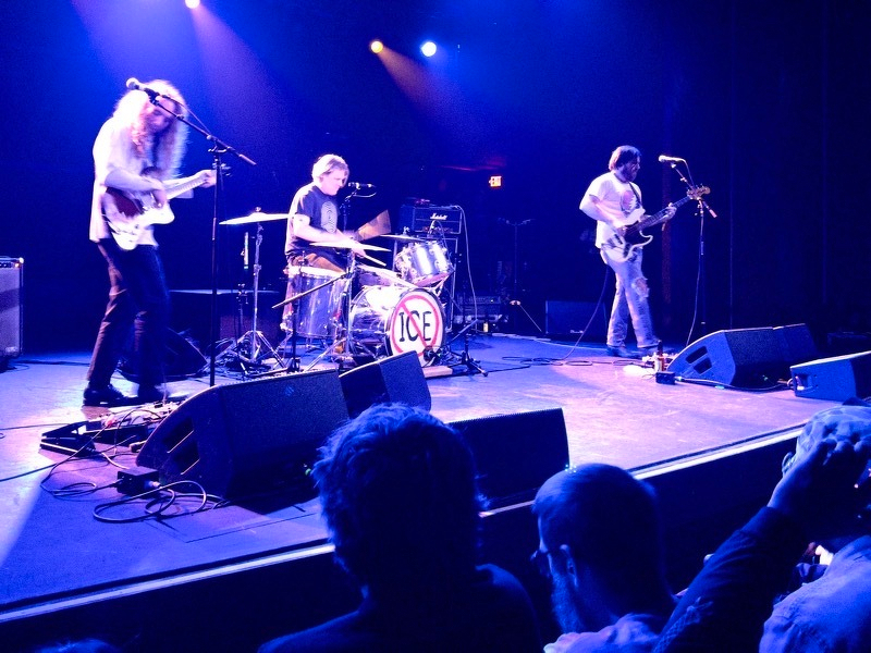 A photo of the band fuzz playing on stage. The drummer's kick drum says "ICE"with a red line crossing it out.