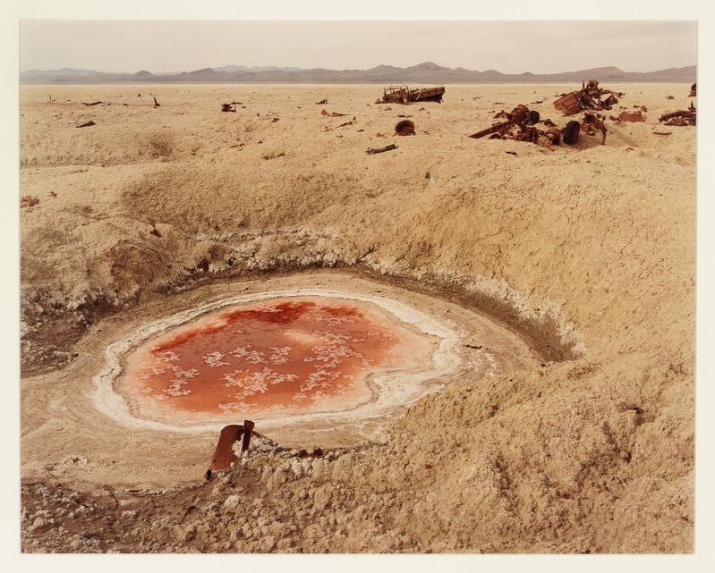 A photo by Richard Misrach showing a bomb crater in the Nevada desert surrounded by broken machinery and filled with a nauseating red liquid