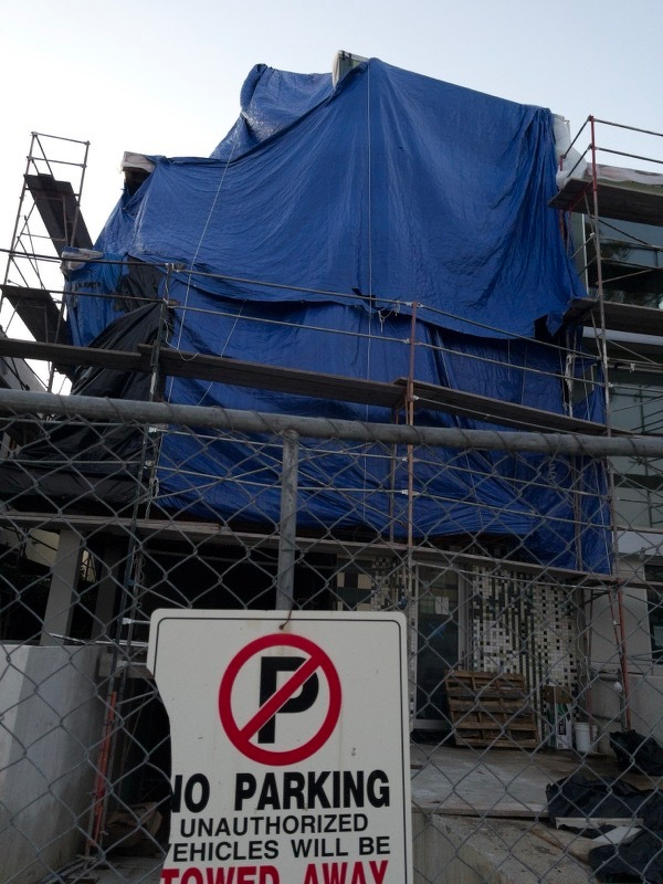 A photo of a structure wreathed in scaffolding and covered in a giant blue tarp. In the foreground you can see a no parking sign wired onto a chainlink fence.