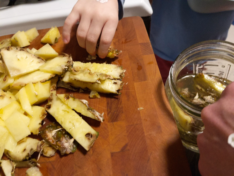 A photo of some cut up pineapple skins on a cutting board with a child's hand grabbing one and putting it in a glass jar