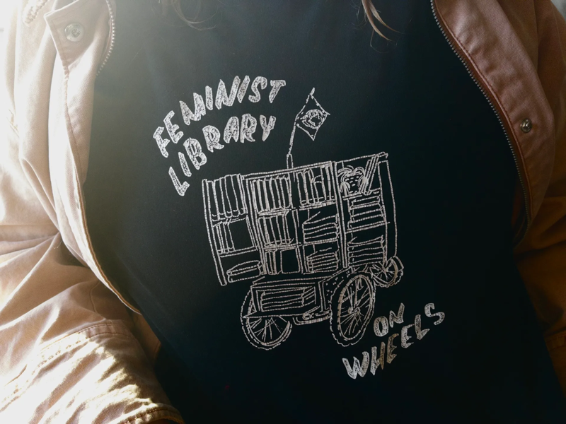 A photo of a woman wearing a t-shirt that says "Feminist Library on Wheels" and shows a line drawing of a little cart that opens up to be tiny bookshelves.