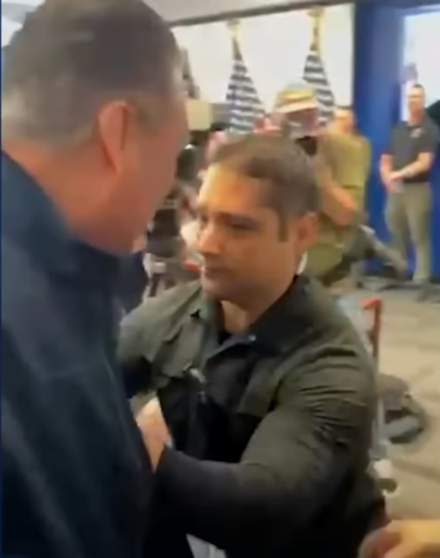 A still frame from a video showing a security guard pushing Senator Alex Padilla away from a podium. The man has a gentle smirk on his face.