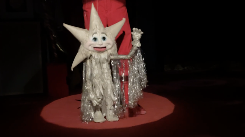 A photo of a marionette star with the puppeteer standing behind, wearing red. The star is encircled by a spotlight.
