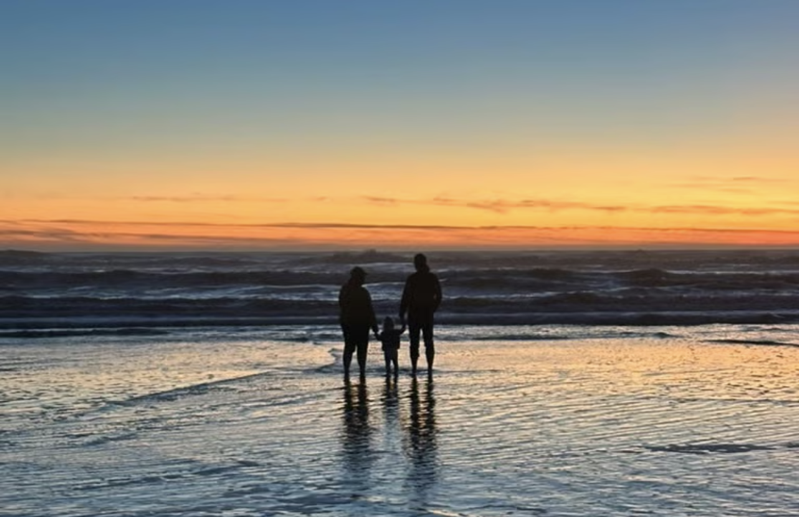 A photo of two adults and a child silhouetted against a sunset. They are standing in the water, which covers their ankles.