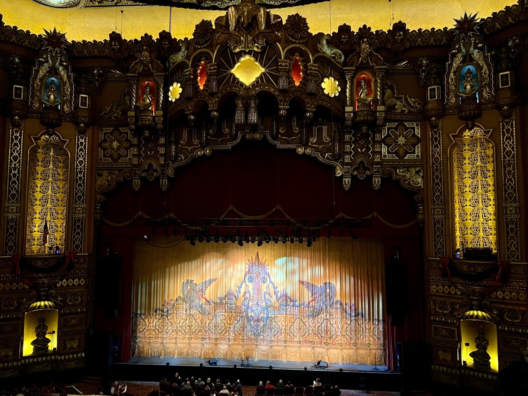 An ornate theater interior with an audience facing a stage showcasing a closed curtain featuring intricate designs, under elaborate architectural decorations and illuminated panels.