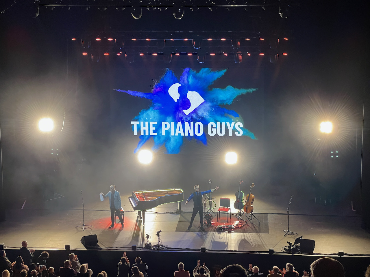 Two musicians on stage with instruments, presenting under a large sign that reads 'THE PIANO GUYS,' with a vibrant blue splash graphic, audience clapping, bright stage lights.