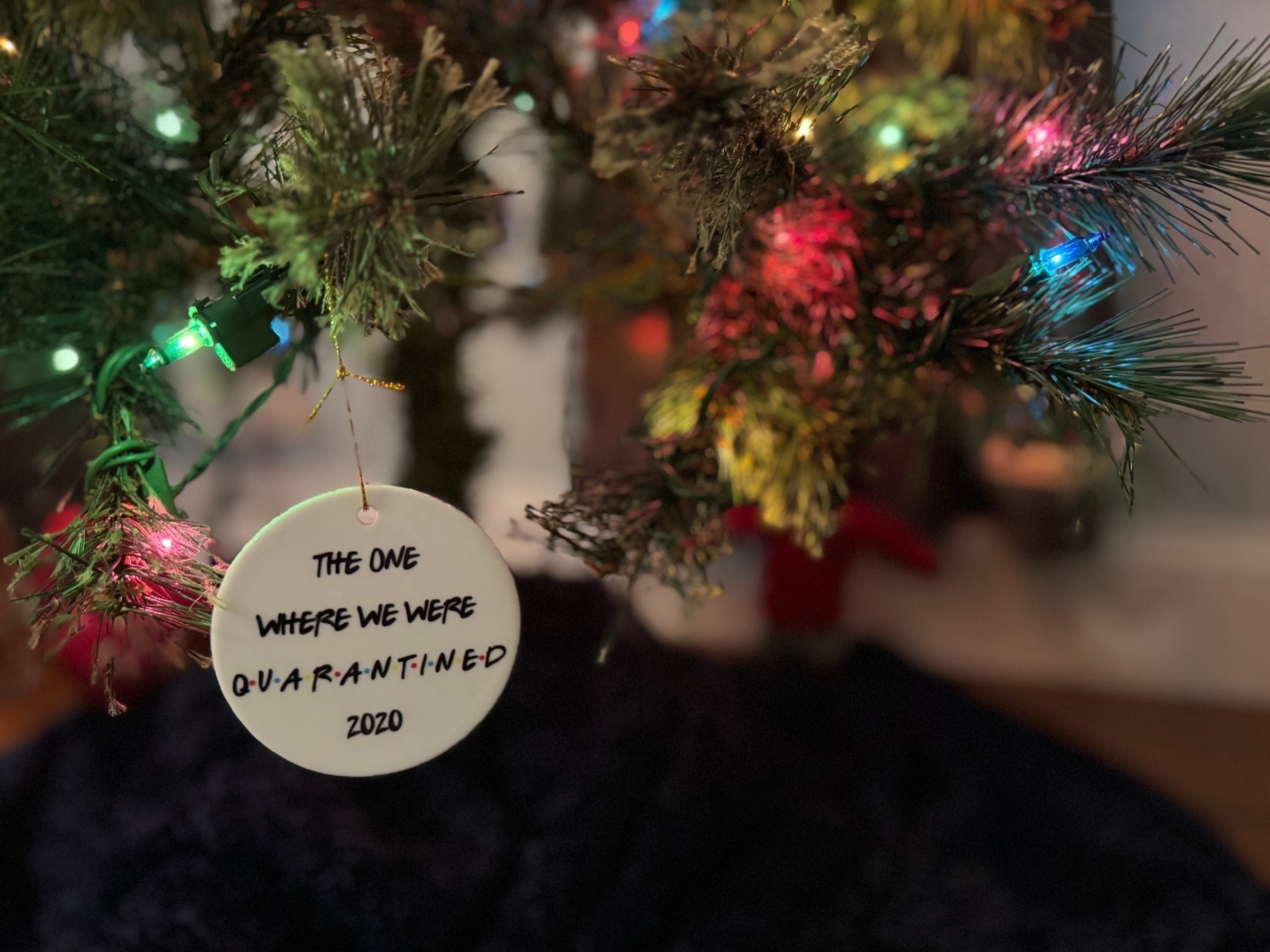 Christmas tree ornament, with colored lights in the background, hanging from the tree. The ornament is round and white, with the following verbiage in the Friends (the TV show) font: 'The one where we were all quarantined, 2020'