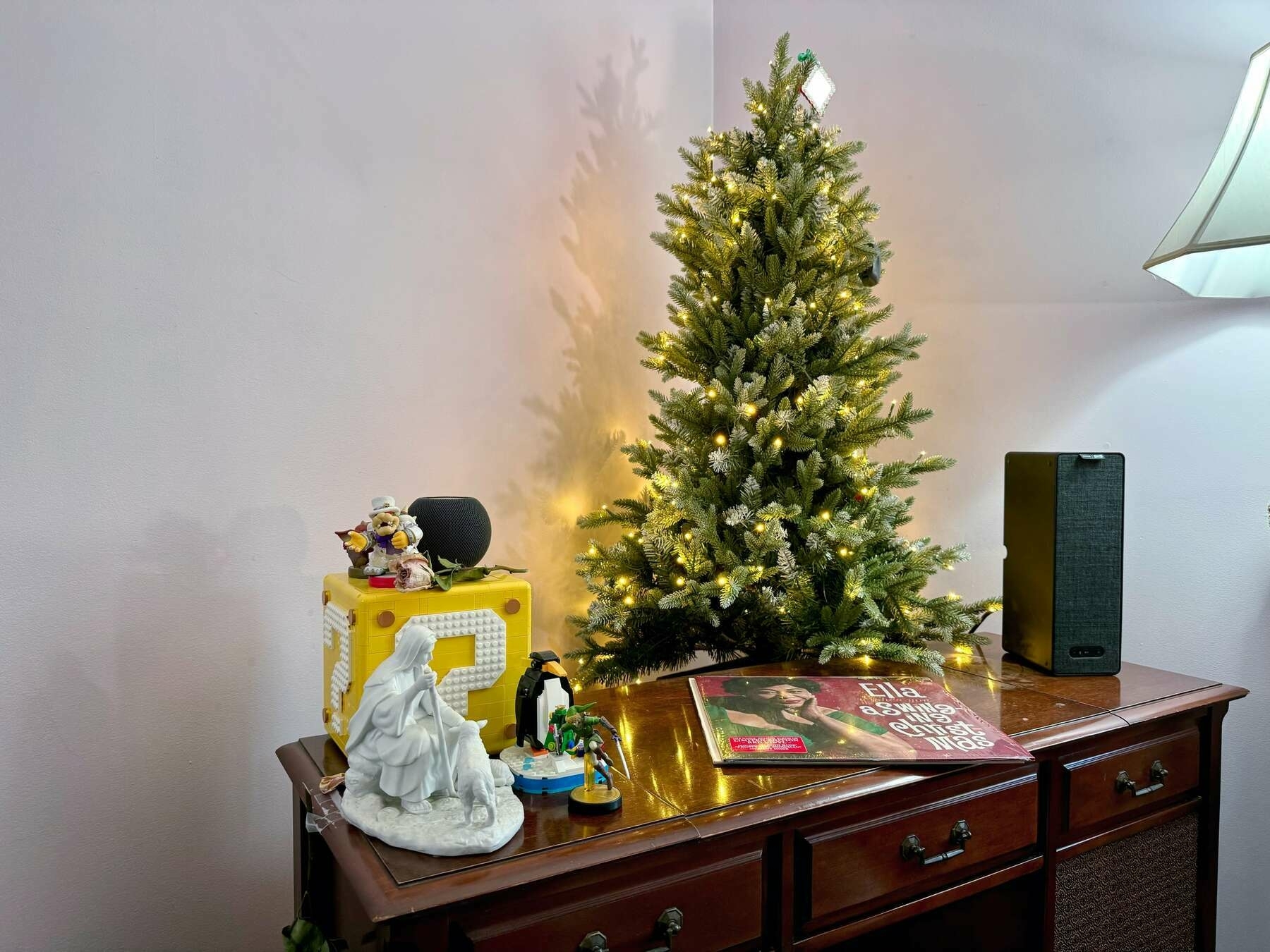 Christmas tree with lights and one ornament at the top (a photo frame with no photo for privacy purposes), in front of a record player, with the end of a lamp on the right side of the photo. An IKEA Sonos speaker is on top of the record player, with an 'Ella' record in the middle and a series of objects is on the left side of the record player: three Nintendo amiibo figures (Link, Bowser, and Bokoblin), a Lego penguin, a Jesus stature, and a N64 Lego cube. On top of the cube is a HomePod mini, along with two of the amiibo, and a dried pink rose.