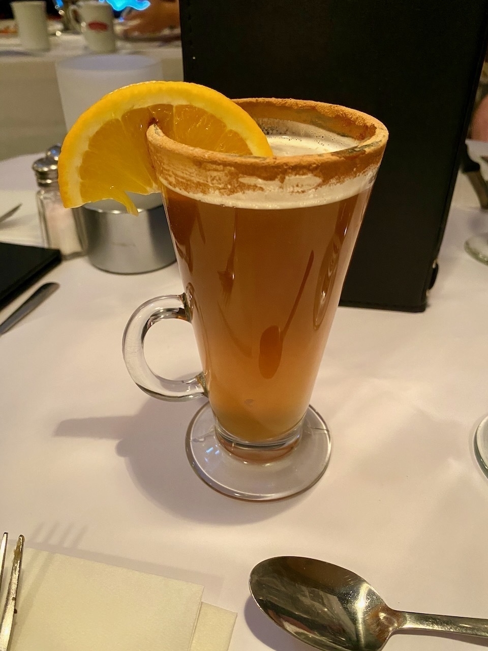 Cider in a glass, the rim dusted with cinnamon powder, with a vertical orange slice on the lip of the glass, on a white-cloth covered table with a spoon in the foreground.