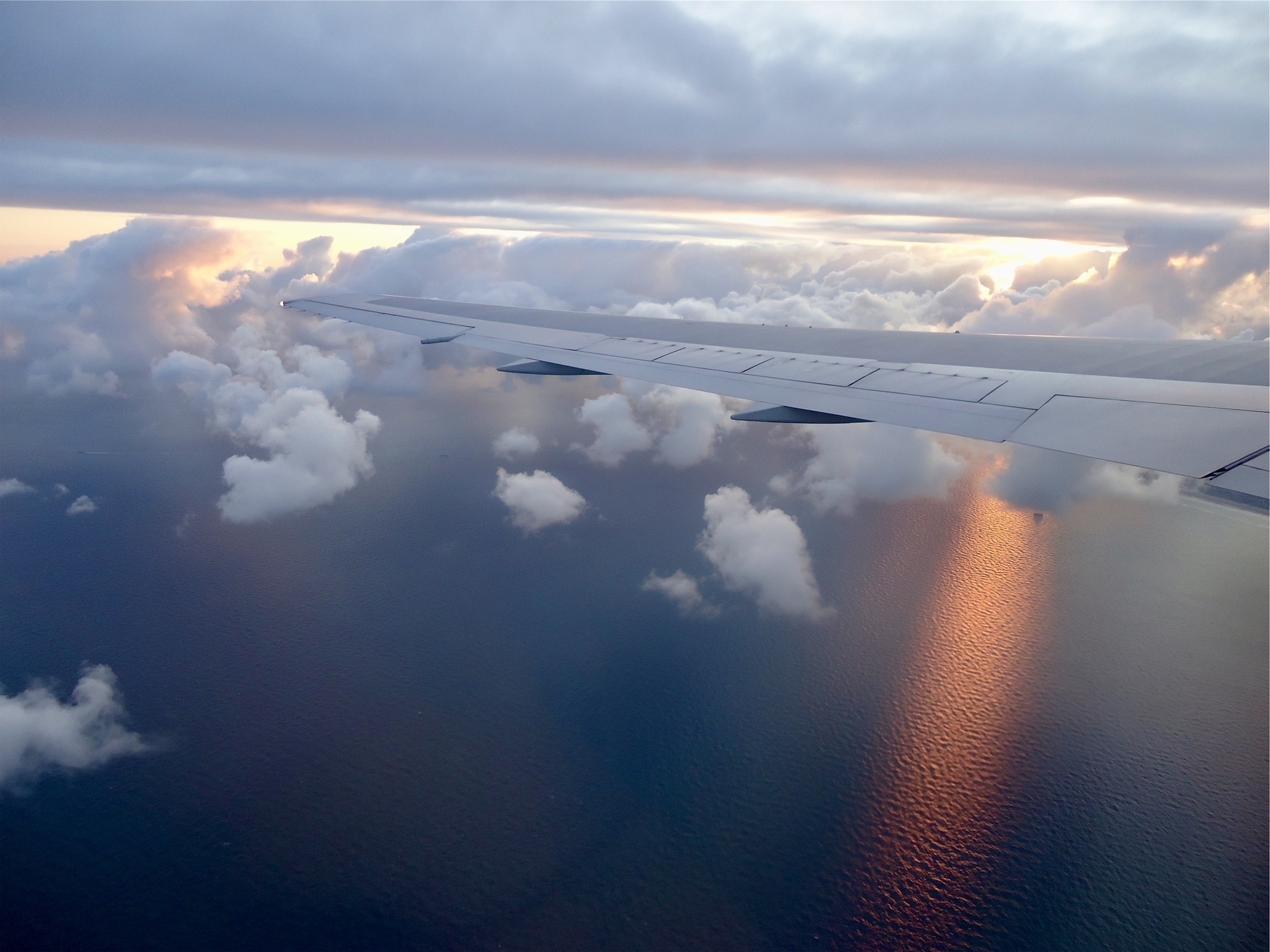 View of the sky and ocean, from an airplane; the ocean is a deep blue/purple below, with a hint of sunlight appearing on the water from behind the clouds, while the white wing of the plan is across the middle of the image.