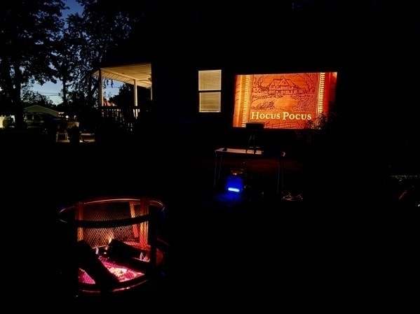 The movie 'Hocus Pocus' projected up on the side of a house at night, frozen at the title screen of the movie for the picture. In the lower-left corner of the image, a glowing fire, and in the upper-left of the image, the fading light of the setting sun.