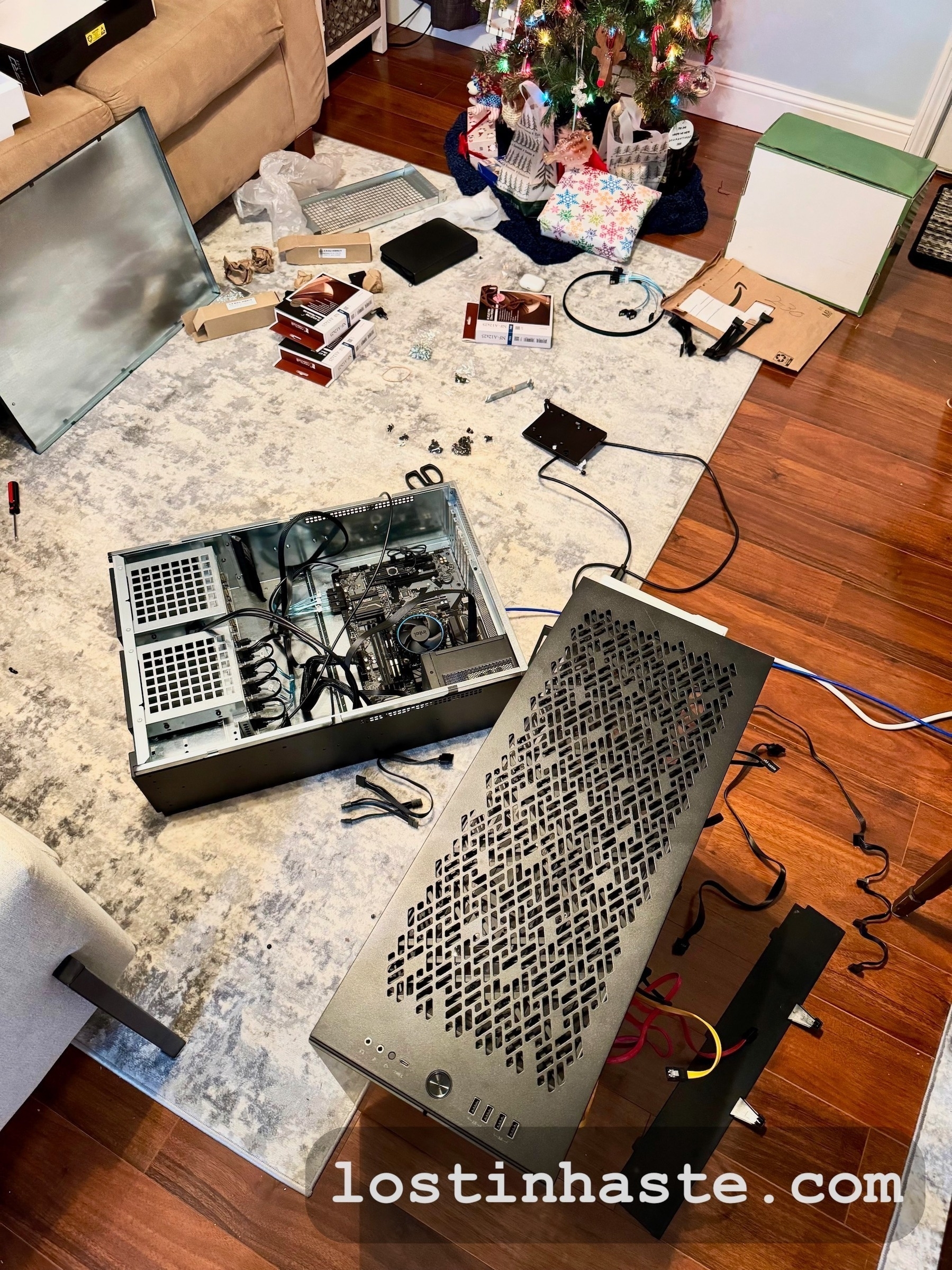 Photo of a tech mess: two computer cases (one horizontal on the floor with open wires exposed and the other vertical with wires poking out), parts on the floor, three computer fan boxes, screws everywhere...on top of a grey/white carpet (the carpet is on top of a hard-wood floor), with a Christmas tree in the background (presents under it), and a couch on the edge of the upper-right side of the photo.