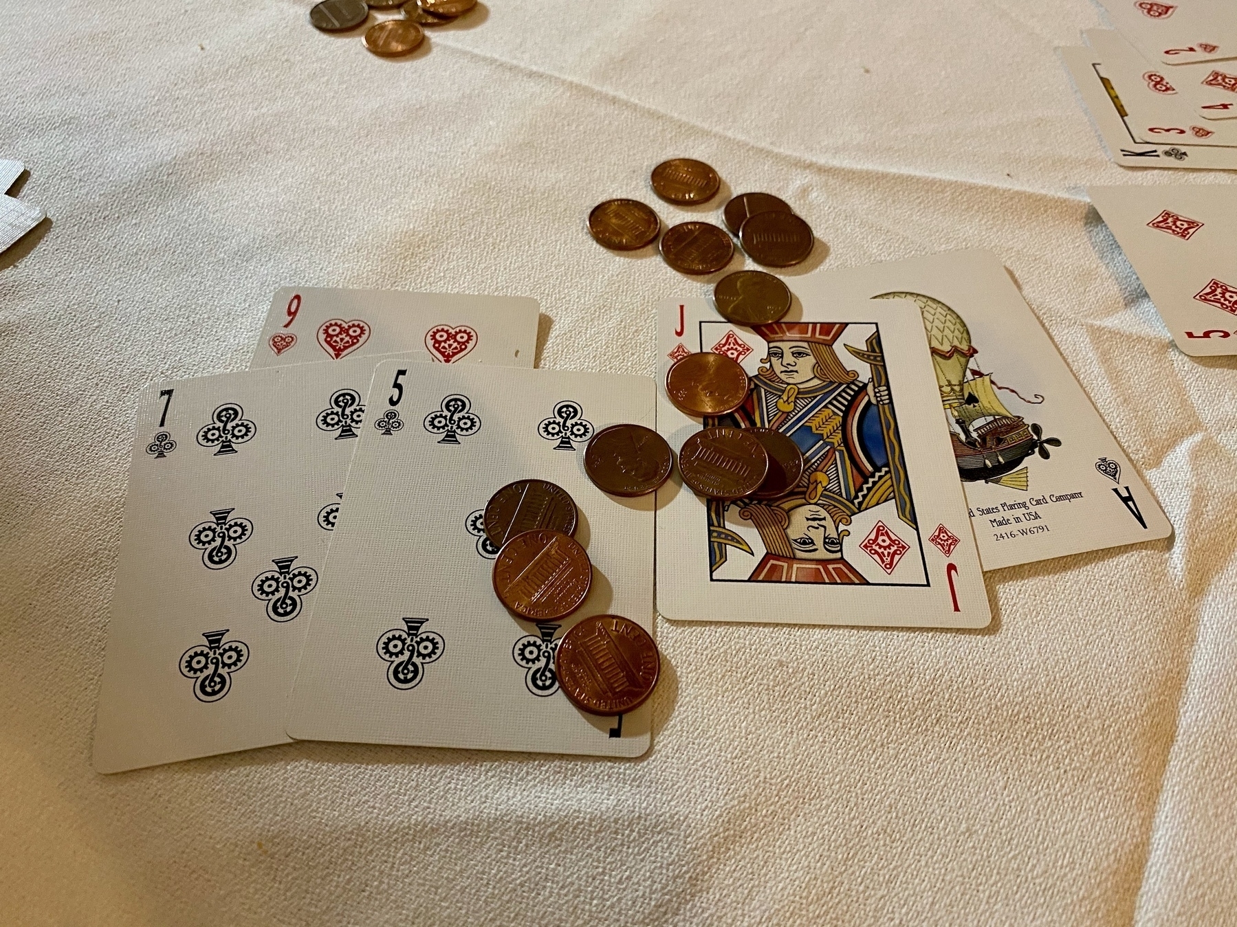 Cards (9 of hearts, 7 and 5 of clubs on one side, with the Jack of diamonds and the Ace of spades on the other side) on a white tablecloth, with pennies on top of them.