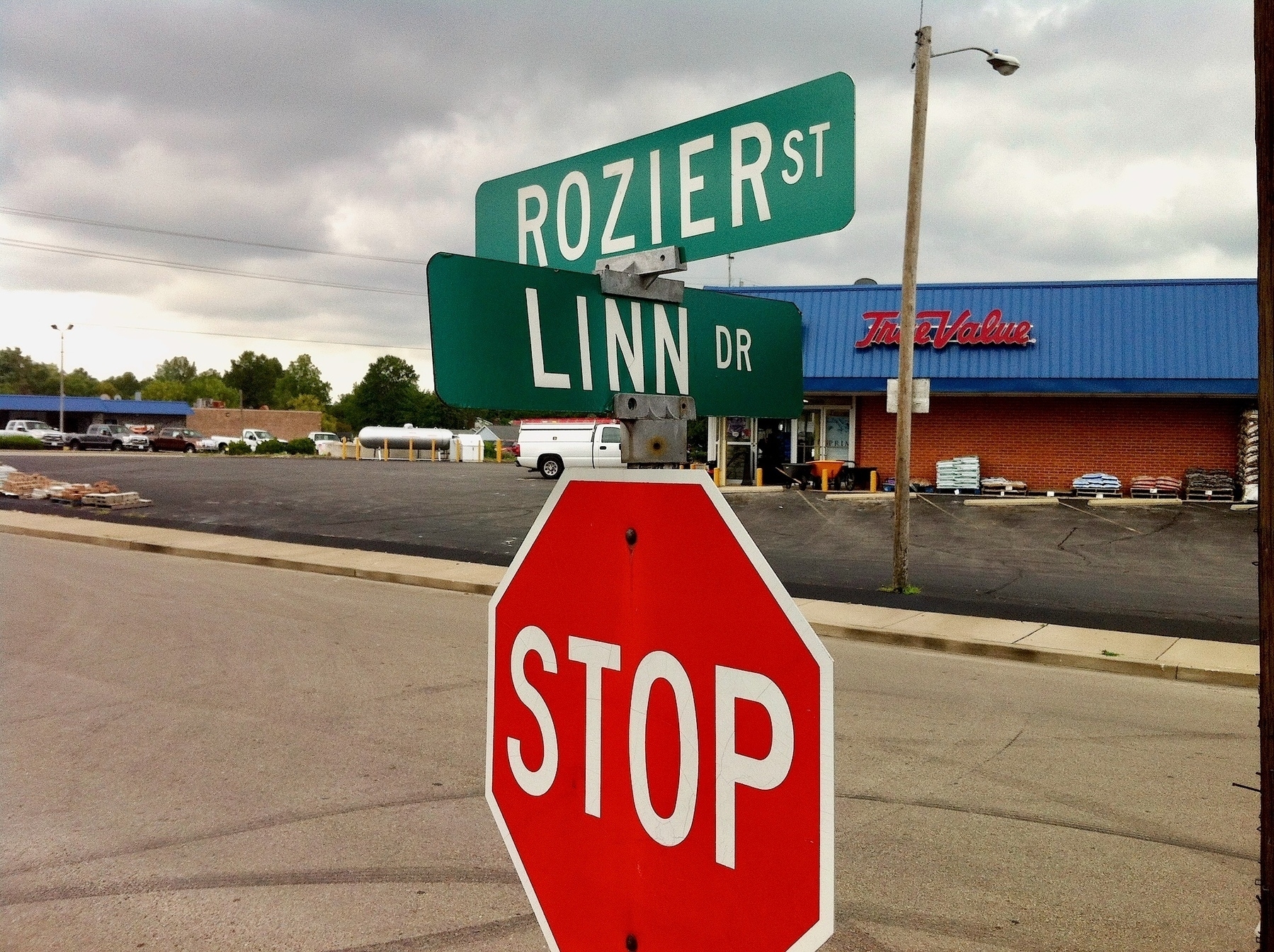 Red stop sign in the foreground, with road names mounted on top: 'Rozier St' and 'Linn Dr'; a True Value hardware store is in the background, the parking lot right behind the sign.