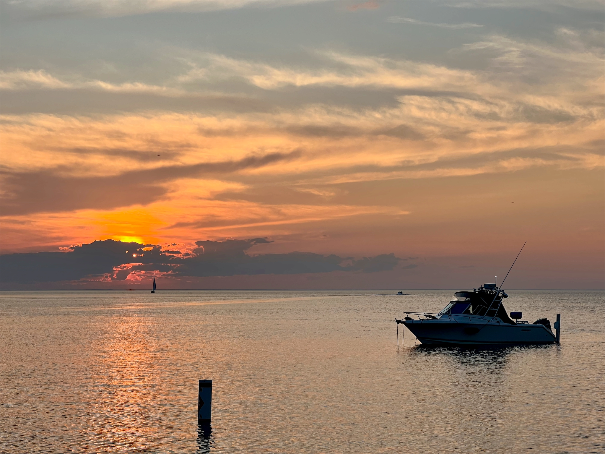 Sunset on one of the Great Lakes (the sun covered by clouds, casting a yellow-orange glow across the sky), with a sailboat in the distance and a fishing boat in the forground.