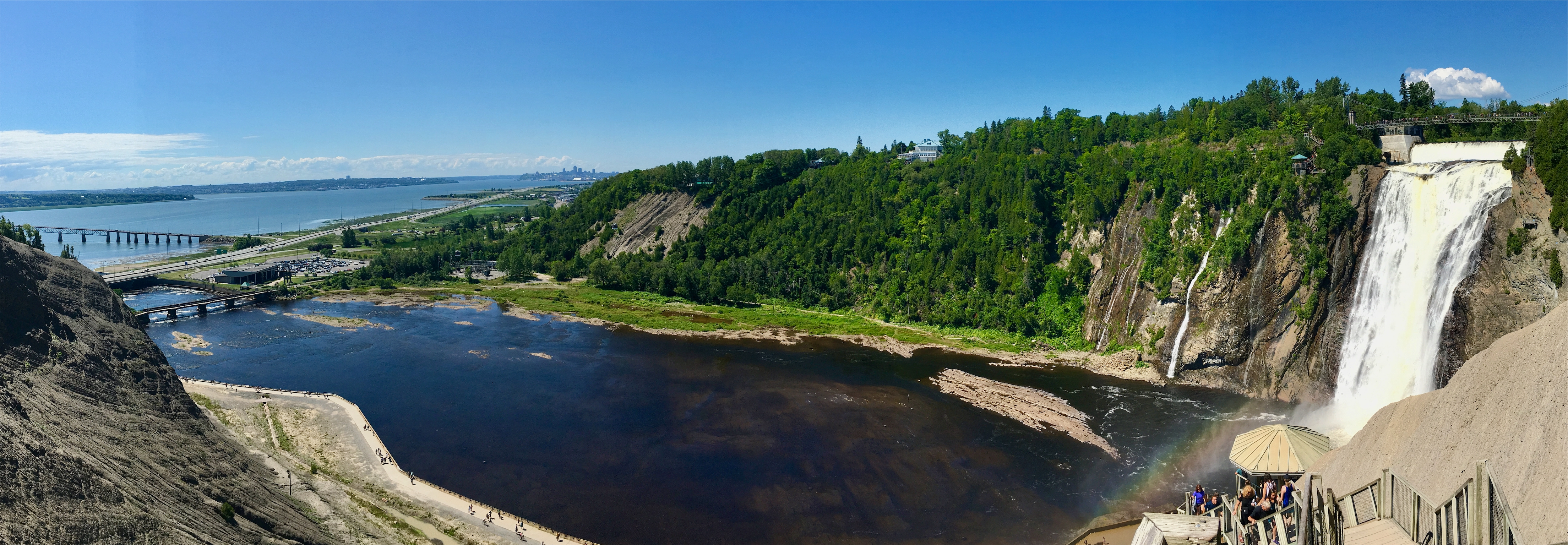 Panoramic view of a water fall on the right, with a bridge going over the falls, wooden steps in the lower-right of the photo, leading down to a covered octagon pavilion on a hill-side, and a large body of water in the center of the photo, flowing out to a river in the distance, in the upper-left of the photo, where several bridges can be seen.
