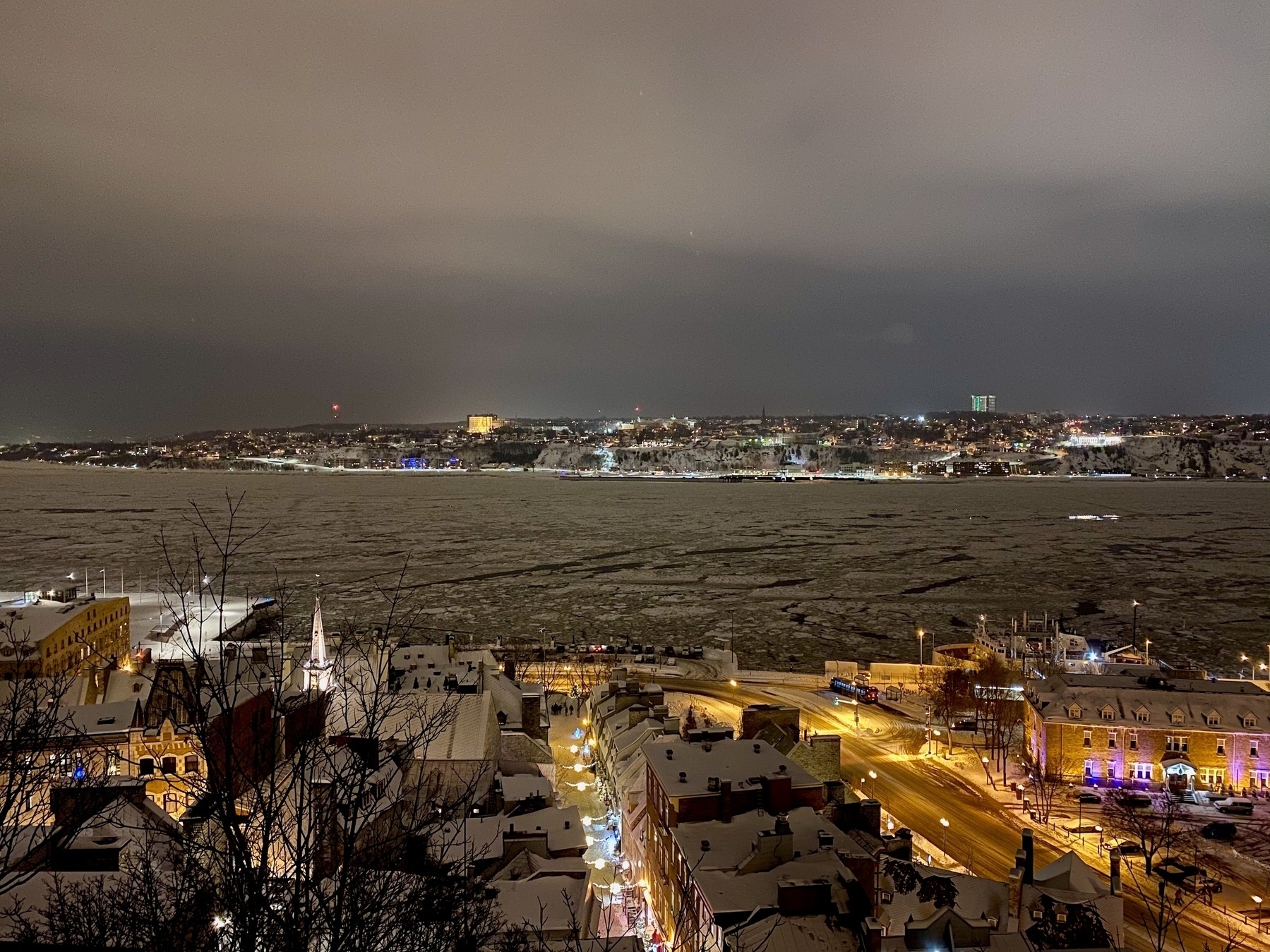 View of the St. Lawrence River, with ice chunks occupying most of the surface, at night, with houses covered in snow and streets with lights at the bottom of the picture. On the upper part of the picture, across the river, lights can be seen up and down a bluff, with a few well-light buildings prominent in the cloudy night sky.