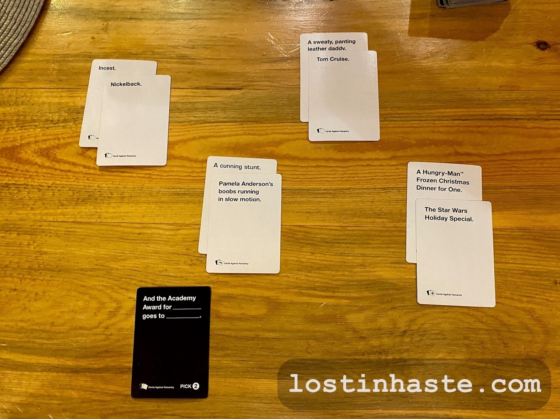 Playing cards are displayed on a wooden table, part of a card game setup with text on them to be combined humorously. Text on cards: 'Incest', 'Nickelback', 'A sweaty, panting leather daddy', 'Tom Cruise', 'A cunning stunt', 'Pamela Andersons boobs running in slow motion', 'A Hungry-Man® Frozen Christmas Dinner for One', 'The Star Wars Holiday Special', 'And the Academy Award for _____ goes to _____ (Black card)', 'PICK 2 (on the black card)'. The image has a 'lostinhaste.com' watermark.