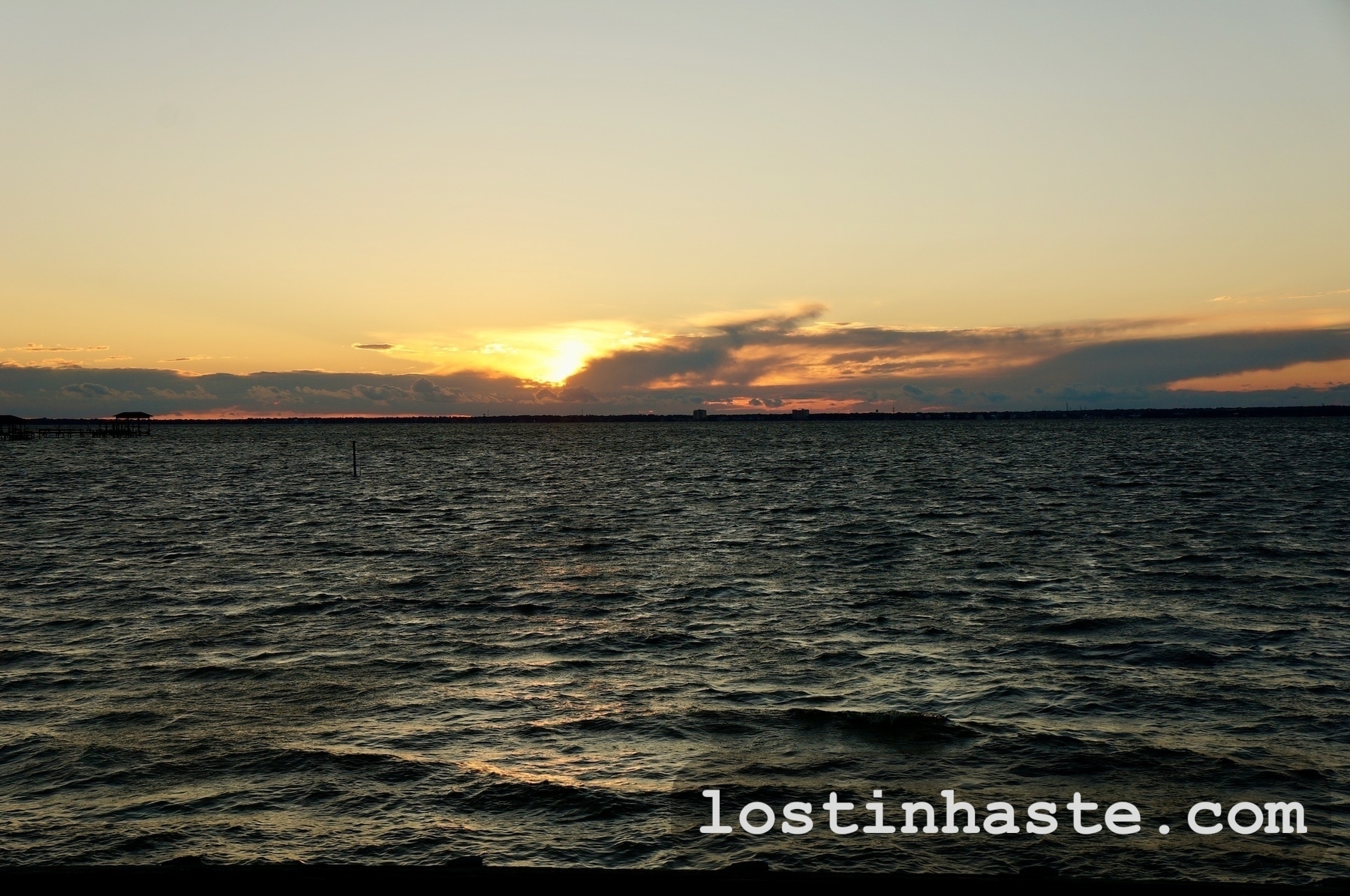 Sunset over choppy water with clouds partly obscuring the sun and darkening the skyline; the text reads 'lostininhaste.com.'