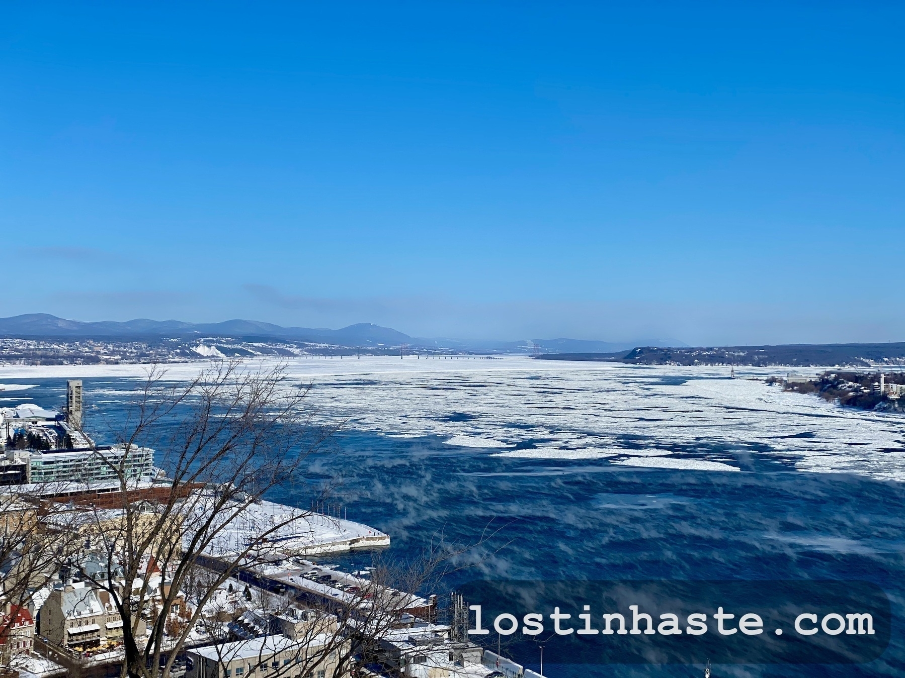 A frozen river flows through a cityscape against a backdrop of mountains under a clear blue sky. Text: lostinhaste.com.