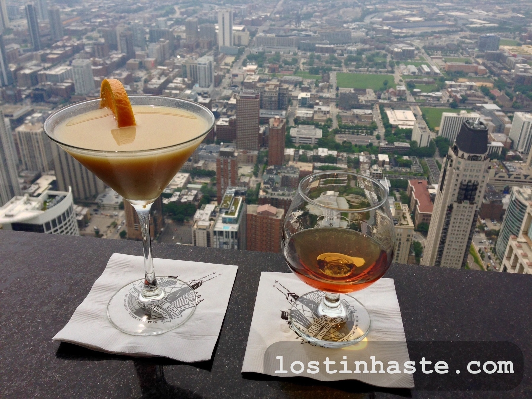 Two drinks on napkins overlook a dense cityscape from a high vantage point. Text: 'lostinhaste.com'
