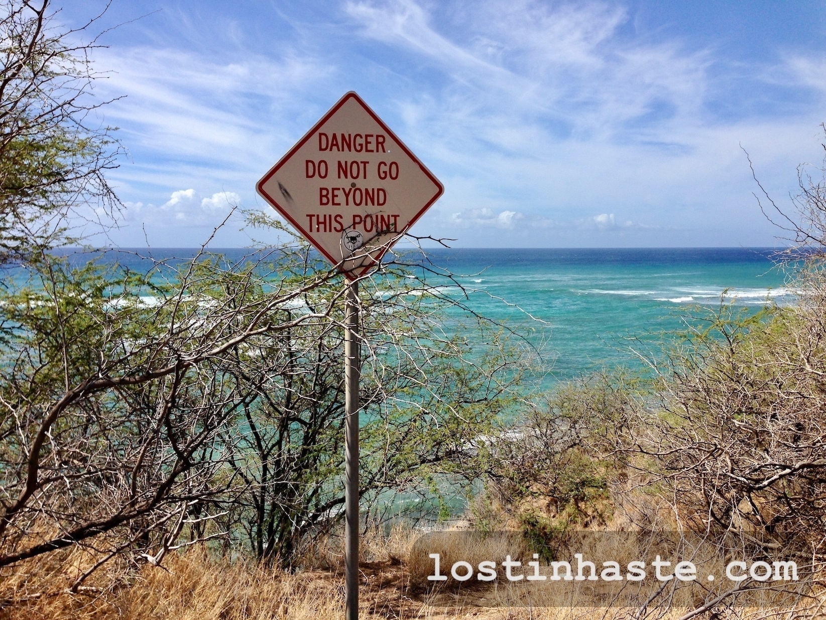 A warning sign reads 'DANGER DO NOT GO BEYOND THIS POINT' against a backdrop of tropical sea and sky. The website address 'lostinmhaste.com' is visible.