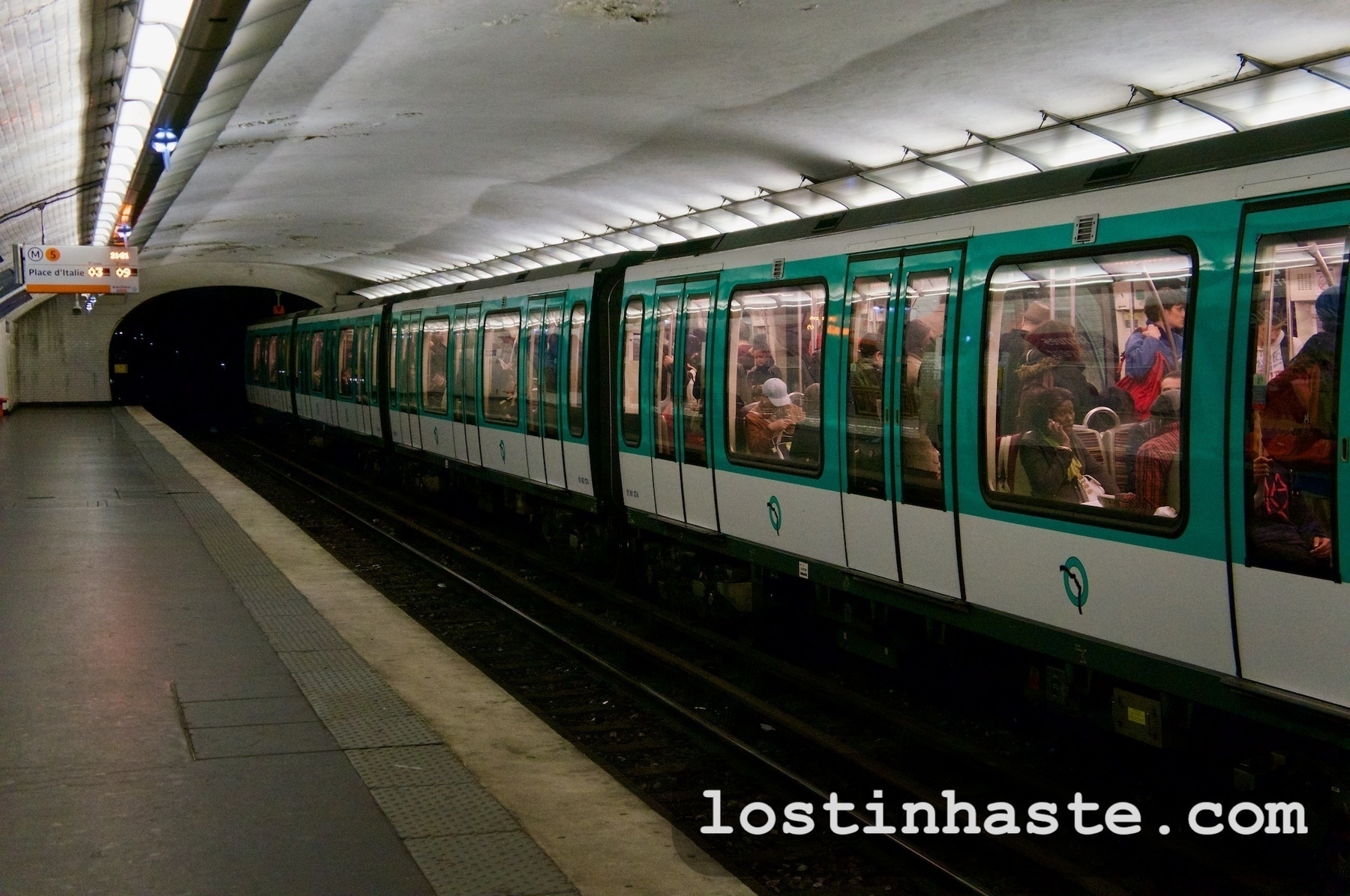 A green metro train is stationary at an underground platform with passengers visible through windows, under a curved white ceiling with lights. The text 'lostinhaste.com' is overlaid at the bottom.