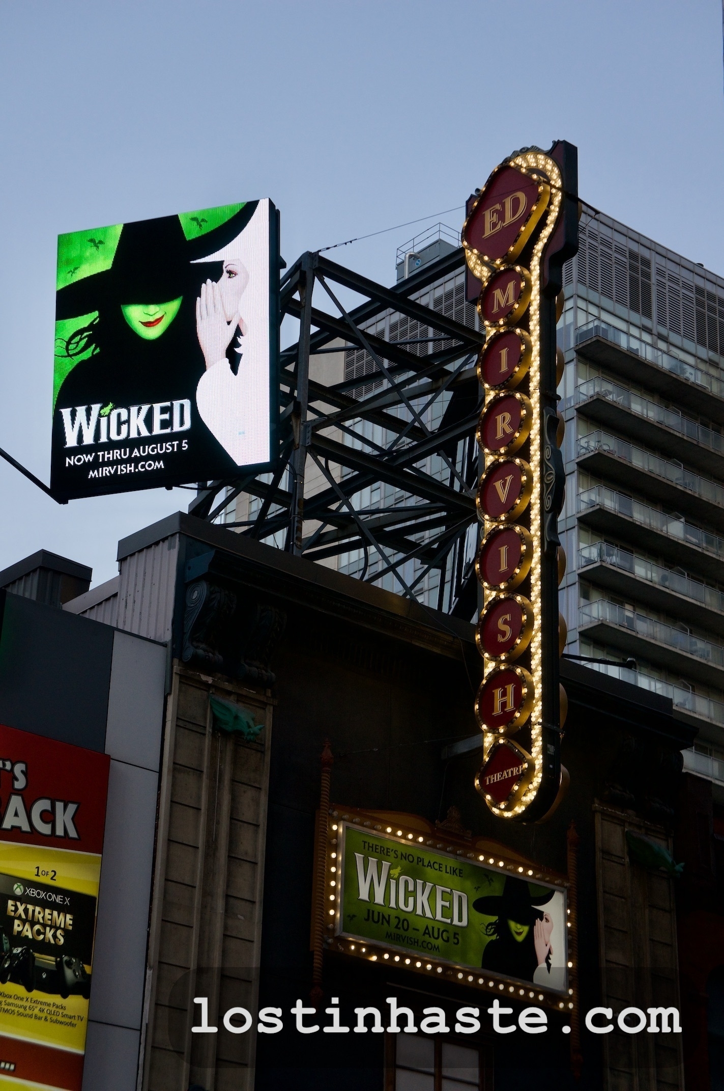 Digital billboard and theater marquee promote the musical 'Wicked' against an urban background; the URL 'mirvish.com' and dates 'NOW THRU August 5' are visible. Lower corner watermark reads 'lostinahaste.com'.