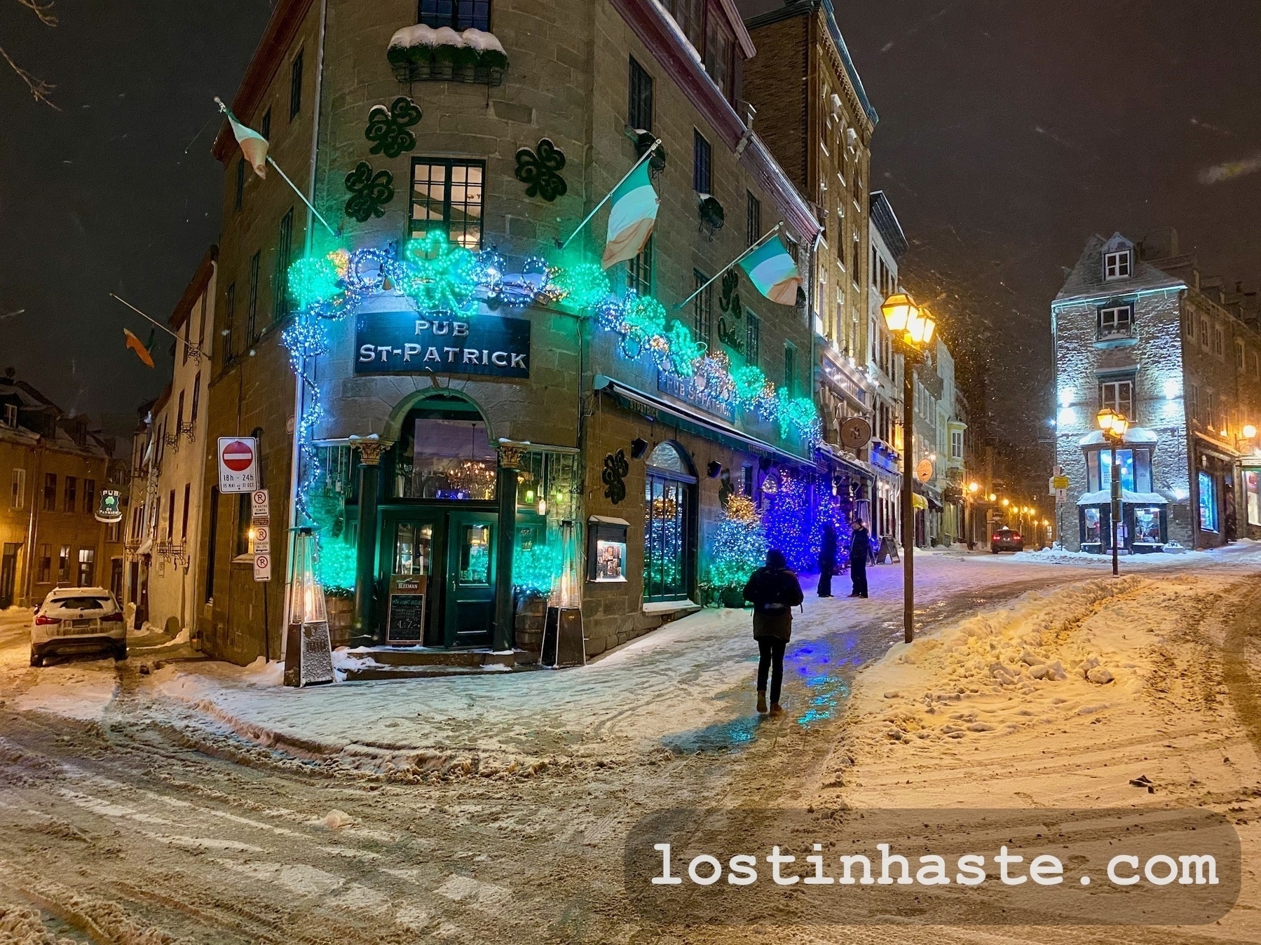 Corner building adorned with blue lights and shamrocks; pedestrians walk snowy streets at night. Text: lostinhaste.com'