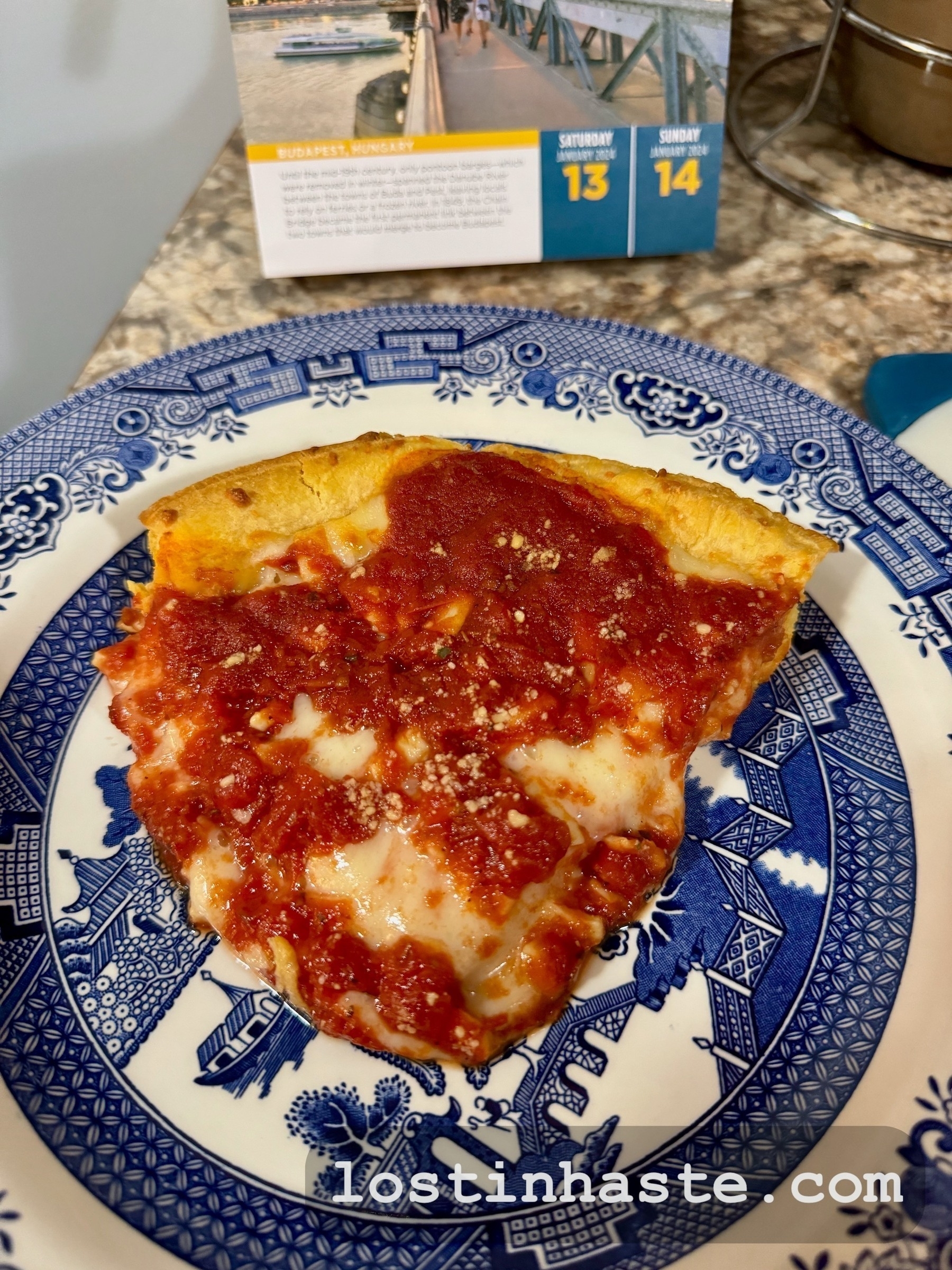 A slice of cheese pizza rests on a blue-and-white patterned plate, with a granular topping, possibly Parmesan, on a kitchen counter with a calendar in the background.